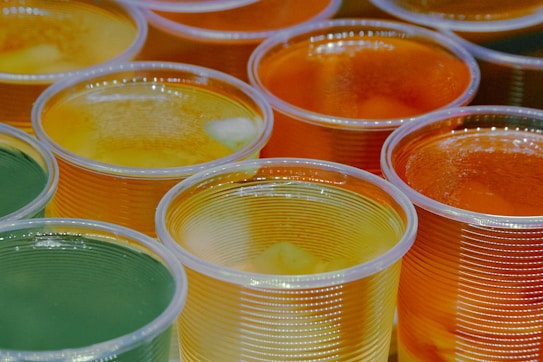 A close-up of several clear plastic cups filled with various colors and types of gelatin. The cups contain green, yellow, and orange gelatin, with some pieces of fruit visible inside. The arrangement of the cups creates a colorful and inviting display.