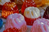 Assorted jelly molds with a glossy texture are arranged on a surface. The molds are in various colors, including pink, white, and orange, creating a vibrant and playful appearance. The translucent nature of the jellies allows light to enhance their bright hues.