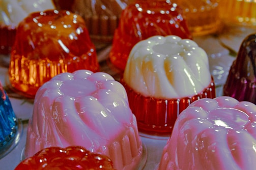 Assorted jelly molds with a glossy texture are arranged on a surface. The molds are in various colors, including pink, white, and orange, creating a vibrant and playful appearance. The translucent nature of the jellies allows light to enhance their bright hues.