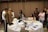 Several people are gathered in a room, seemingly engaged in a meeting or collaborative session. A round table covered with documents, laptops, beverages, and various objects is in the foreground. Some people are sitting around the table working on laptops while others are standing nearby in discussions and looking at a piece of paper one of them is holding up. The room has decorative elements and subdued lighting.