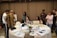 Several people are gathered in a room, seemingly engaged in a meeting or collaborative session. A round table covered with documents, laptops, beverages, and various objects is in the foreground. Some people are sitting around the table working on laptops while others are standing nearby in discussions and looking at a piece of paper one of them is holding up. The room has decorative elements and subdued lighting.