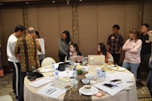 Several people are gathered in a room, seemingly engaged in a meeting or collaborative session. A round table covered with documents, laptops, beverages, and various objects is in the foreground. Some people are sitting around the table working on laptops while others are standing nearby in discussions and looking at a piece of paper one of them is holding up. The room has decorative elements and subdued lighting.
