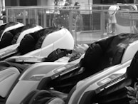 A person is reclining in a line of futuristic massage chairs, located in what appears to be a public space such as a mall. The chairs have a sleek design with visible control panels, and the setting is quiet and relaxed.