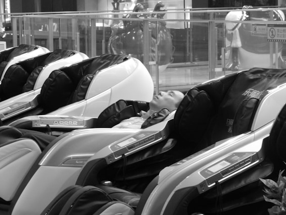 A person is reclining in a line of futuristic massage chairs, located in what appears to be a public space such as a mall. The chairs have a sleek design with visible control panels, and the setting is quiet and relaxed.