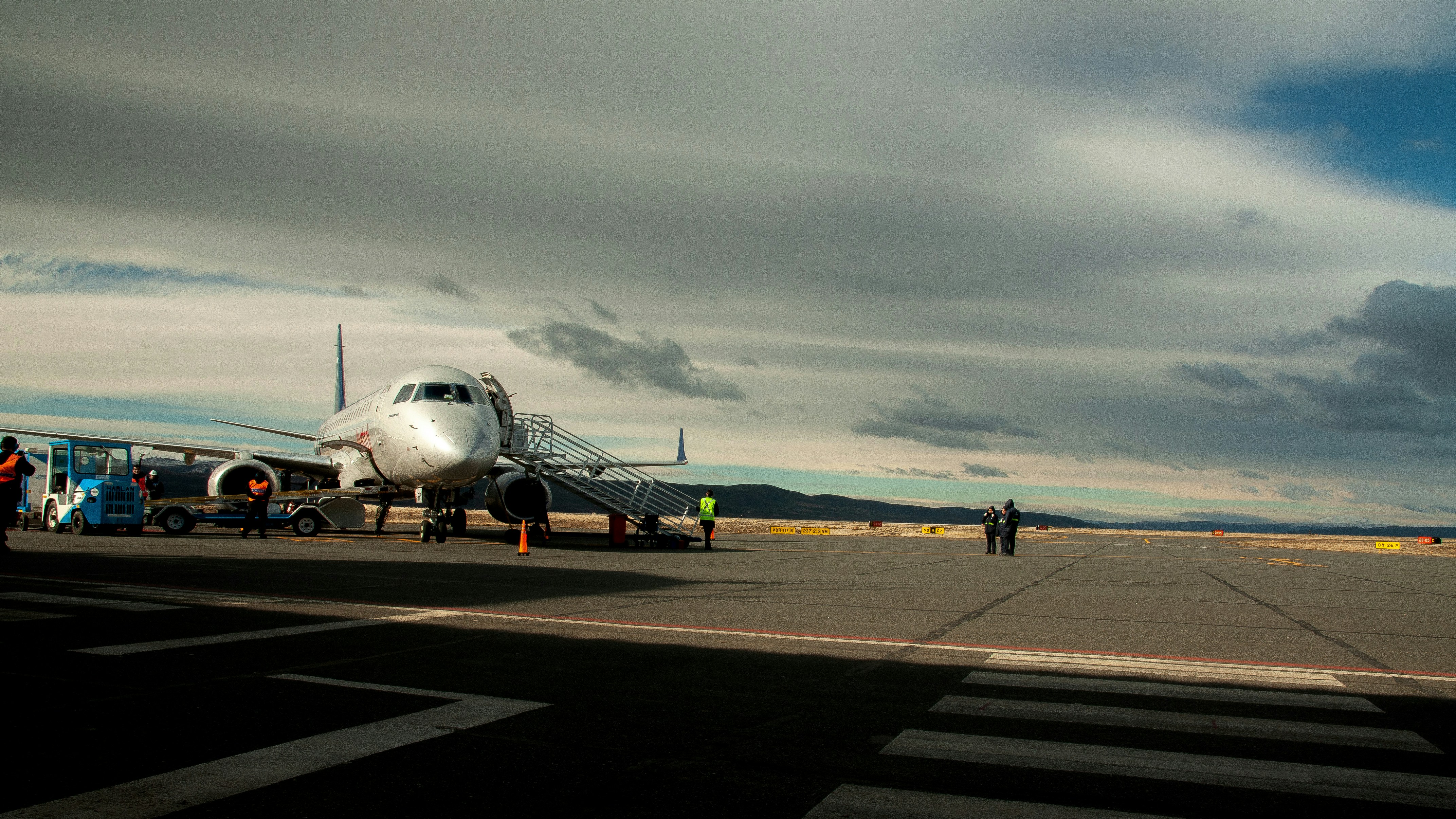 Wide-body commercial airliner at gate, overcast sky, dim industrial airport lighting, dark concrete ramp, atmospheric shadows