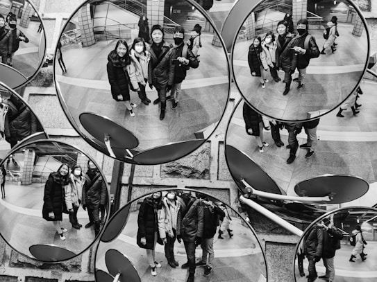 A black and white image displaying several convex mirrors reflecting a group of people standing together in a public space. The people are in winter clothing, and some are wearing masks. The mirrors are mounted on a textured wall and create a kaleidoscopic effect with multiple reflections of the same scene.