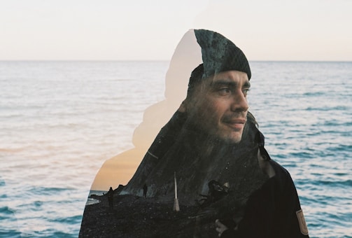 A double exposure photograph blending a serene coastal scene with the portrait of a man wearing a beanie. The ocean provides a tranquil backdrop, and his thoughtful expression adds a reflective quality to the composition.