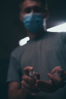 A person wearing a blue face mask stands in a dimly lit environment, holding a small spray bottle in their hands. The focus is on the hands and the bottle, with the rest of the image blurred, giving it a mysterious and moody atmosphere.