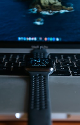 A smartwatch with a perforated strap is placed on top of a laptop keyboard. The digital display of the watch shows the time and other information. The laptop screen in the background displays a desktop with a nature-themed wallpaper and a dock of icons at the bottom.