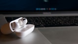 Wireless earbuds with a white case are placed on a silver laptop, with a black keyboard and screen in the background.