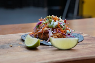 A vibrant taco resting on a wooden board, topped with a mix of colorful shredded vegetables, creamy sauce, and garnished with green herbs. A slice of lime sits nearby, enhancing the dish's appeal.