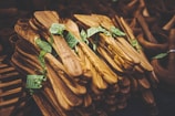 A collection of intricately crafted wooden kitchen utensils, neatly stacked and adorned with green tags. The utensils display rich wood grain patterns with a natural, polished finish.
