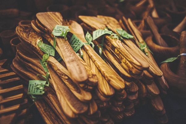 A collection of intricately crafted wooden kitchen utensils, neatly stacked and adorned with green tags. The utensils display rich wood grain patterns with a natural, polished finish.