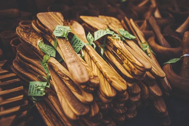 A collection of intricately crafted wooden kitchen utensils, neatly stacked and adorned with green tags. The utensils display rich wood grain patterns with a natural, polished finish.