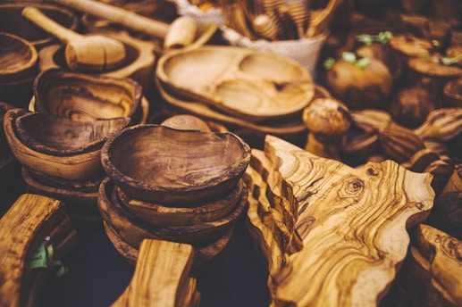 A collection of handcrafted wooden bowls and utensils with rich, textured grain patterns. The items are arranged closely together, showcasing a variety of shapes and sizes. The overall appearance is rustic and artisanal, highlighting the natural beauty of the wood.