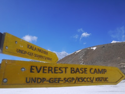 Two yellow directional signs indicate the directions to Kala Patthar and Everest Base Camp against a backdrop of snowy mountain peaks and a clear blue sky.