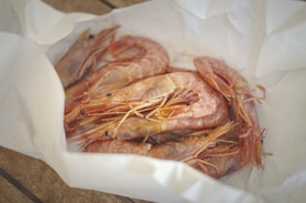 A pile of raw shrimp with long antennae and translucent shells placed on white parchment paper. The shrimp have a pinkish hue, and the surface of the paper gives a clean, fresh appearance.