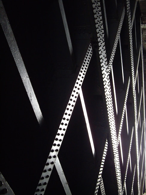A series of metallic beams with a grid-like pattern creating an architectural structure. The beams intersect at angles, casting shadows and reflecting light, forming a complex geometric design.