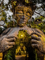 A large wooden sculpture of a humanoid figure with intricate carvings stands amid lush greenery. The figure's hands appear to be drawing open the chest, revealing a passage adorned with hanging plants and a warm, glowing light. There are two signs attached to the trees on either side, reading 'Raw Love Cafe' and 'Ahau Tulum.'
