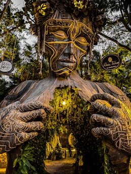 A large wooden sculpture of a humanoid figure with intricate carvings stands amid lush greenery. The figure's hands appear to be drawing open the chest, revealing a passage adorned with hanging plants and a warm, glowing light. There are two signs attached to the trees on either side, reading 'Raw Love Cafe' and 'Ahau Tulum.'