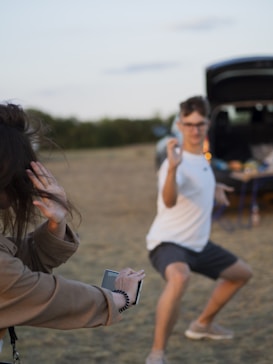 A person in a playful pose while another holds a smartphone toward them, suggesting they might be recording or taking a photo. The scene is set outdoors, with a blurred background of grass and trees.