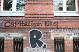 A brick wall of a building is covered with various graffiti and stencils. The prominent graffiti reads 'Capitalism Kills' in large black letters. Below, there are smaller stencils with phrases like 'Forward to Feminism' and 'Besetzen statt kapitulieren'. The wall has a set of barred windows with grills, and there are multiple layers of colorful spray paint tags scattered across the surface.