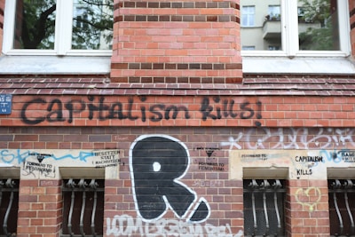 A brick wall of a building is covered with various graffiti and stencils. The prominent graffiti reads 'Capitalism Kills' in large black letters. Below, there are smaller stencils with phrases like 'Forward to Feminism' and 'Besetzen statt kapitulieren'. The wall has a set of barred windows with grills, and there are multiple layers of colorful spray paint tags scattered across the surface.