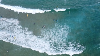 Aerial view of a group of people surfing in the ocean. The waves are white and foamy, contrasting with the darker blue-green water. Some surfers are riding waves while others are waiting for the next set. The scene is vibrant and dynamic, capturing the energy of the ocean and the thrill of surfing.
