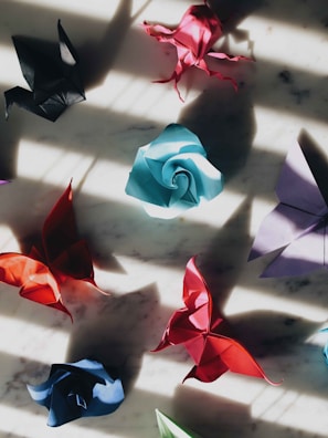 A collection of colorful origami figures is scattered across a marble surface, illuminated by dramatic shadows. The origami pieces include a black crane, a red butterfly, a blue rose, and several other shapes, primarily in shades of red, blue, and purple.