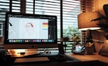A cozy workspace with a desktop computer displaying graphic design software, showing a cartoon character. The desk is illuminated by a warm table lamp and features various gadgets, potted plants, and a stylized digital display with the time. Large windows with blinds offer a view of foliage outside, creating a relaxed atmosphere.
