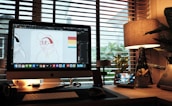 A cozy workspace with a desktop computer displaying graphic design software, showing a cartoon character. The desk is illuminated by a warm table lamp and features various gadgets, potted plants, and a stylized digital display with the time. Large windows with blinds offer a view of foliage outside, creating a relaxed atmosphere.
