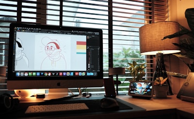 A cozy workspace with a desktop computer displaying graphic design software, showing a cartoon character. The desk is illuminated by a warm table lamp and features various gadgets, potted plants, and a stylized digital display with the time. Large windows with blinds offer a view of foliage outside, creating a relaxed atmosphere.