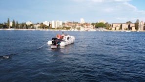 A small inflatable boat with an outboard motor is floating on the water, occupied by several people. In the background, a cityscape with residential buildings, trees, and a clear blue sky is visible. The water appears calm, reflecting the surrounding scenery.