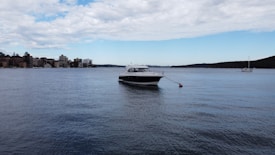 A motorboat is anchored on calm water with a buoy tethered to its bow. In the background, there are buildings lined along the shore under a partly cloudy sky. Another sailboat is visible further in the distance, and the landscape features gentle, rolling hills on the right.