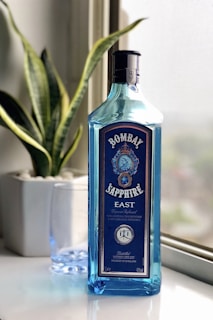A blue bottle of Bombay Sapphire East gin is placed on a white surface near a window. The bottle is tall with a distinct blue hue, featuring intricate labels and branding. Behind the bottle is a green houseplant in a white pot, and next to it is a clear glass.