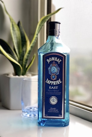 A blue bottle of Bombay Sapphire East gin is placed on a white surface near a window. The bottle is tall with a distinct blue hue, featuring intricate labels and branding. Behind the bottle is a green houseplant in a white pot, and next to it is a clear glass.
