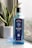 A blue bottle of Bombay Sapphire East gin is placed on a white surface near a window. The bottle is tall with a distinct blue hue, featuring intricate labels and branding. Behind the bottle is a green houseplant in a white pot, and next to it is a clear glass.