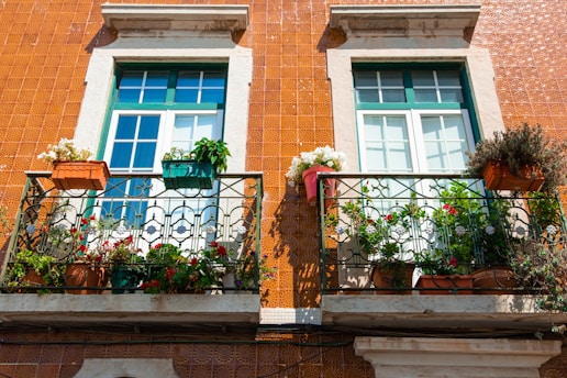 Two windows with white frames and green accents are set in a brown tiled wall. Each window has a small balcony adorned with a variety of potted plants, flowers, and greenery. The balconies have ornate black iron railings, adding a decorative touch.
