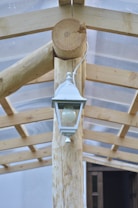 A white outdoor lantern is mounted on a wooden beam structure with a transparent corrugated plastic roof. The design features natural, unfinished wood with distinct grain patterns and a simple, rustic look.