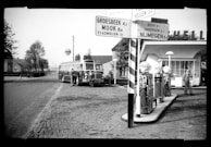 A vintage scene at a gas station with classic fuel pumps and a large bus parked nearby. Two individuals can be seen near the bus, suggesting a moment of rest or travel stopover. The signage in the foreground provides directions to nearby locations, indicating this is a crossroads or travel junction. The architecture and styling suggest mid-20th century.