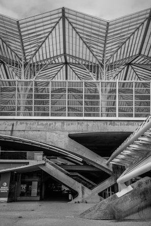 A detailed view of a modern architectural structure with an intricate glass and steel roof design. The geometric patterns created by the intersecting lines provide a visually complex and striking appearance. Below the roof, a concrete base supports the structure with visible diagonal beams adding to the architectural aesthetics.