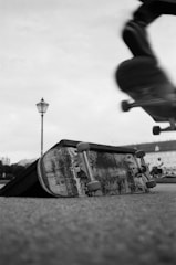 A dynamic scene featuring a skateboard in motion, captured mid-air against a blurred background. On the ground, an abandoned skateboard is positioned on a ramp, displaying signs of wear and tear. A lamp post is visible in the distance, providing a sense of depth to the urban setting.