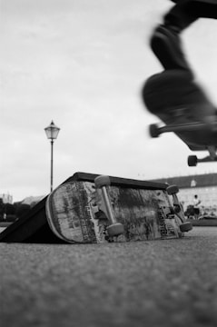 A dynamic scene featuring a skateboard in motion, captured mid-air against a blurred background. On the ground, an abandoned skateboard is positioned on a ramp, displaying signs of wear and tear. A lamp post is visible in the distance, providing a sense of depth to the urban setting.