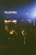 A storefront with the brand name Valentino illuminated against a dark background, at night. Silhouettes of people and objects appear in the shadowy foreground, partially lit by a nearby light source.
