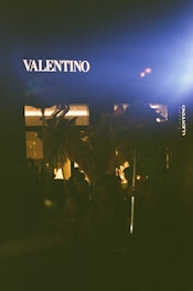 A storefront with the brand name Valentino illuminated against a dark background, at night. Silhouettes of people and objects appear in the shadowy foreground, partially lit by a nearby light source.