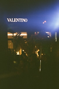 A storefront with the brand name Valentino illuminated against a dark background, at night. Silhouettes of people and objects appear in the shadowy foreground, partially lit by a nearby light source.