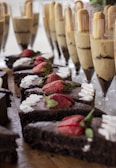 Sliced chocolate cakes topped with fresh strawberries and cream are displayed on a wooden surface. In the background, tall glasses are filled with layered desserts, featuring creamy and chocolate layers topped with cookies.