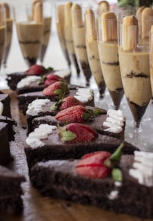 Sliced chocolate cakes topped with fresh strawberries and cream are displayed on a wooden surface. In the background, tall glasses are filled with layered desserts, featuring creamy and chocolate layers topped with cookies.