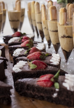 Sliced chocolate cakes topped with fresh strawberries and cream are displayed on a wooden surface. In the background, tall glasses are filled with layered desserts, featuring creamy and chocolate layers topped with cookies.