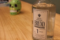 A clear glass bottle of Mezcal Artesanal with a label reading 'BRUXO Elixir de los Terrenales' is placed on a wooden surface. In the background, there is a green decorative skull with black eye sockets and white teeth, slightly out of focus.